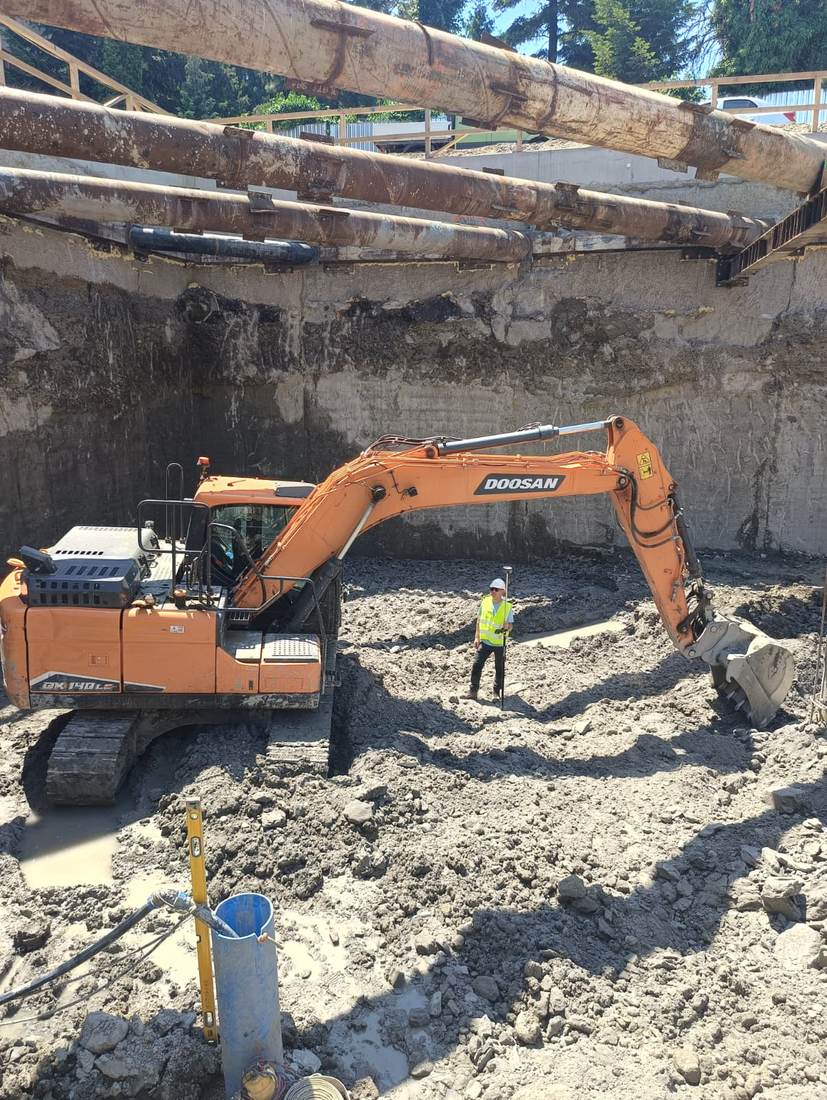 Excavator lucrând într-o groapă de fundație, cu un membru al echipei realizând măsurători de control.