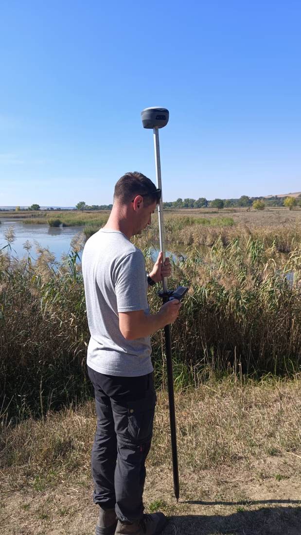 Topograf efectuând măsurători GNSS în teren, cu controller în mână, lângă un iaz.