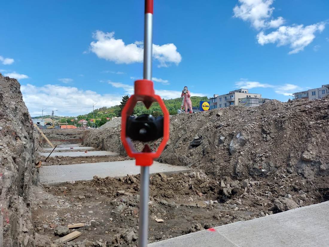 Prismă reflectorizantă în prim-plan, cu un membru al echipei realizând măsurători pe șantier.