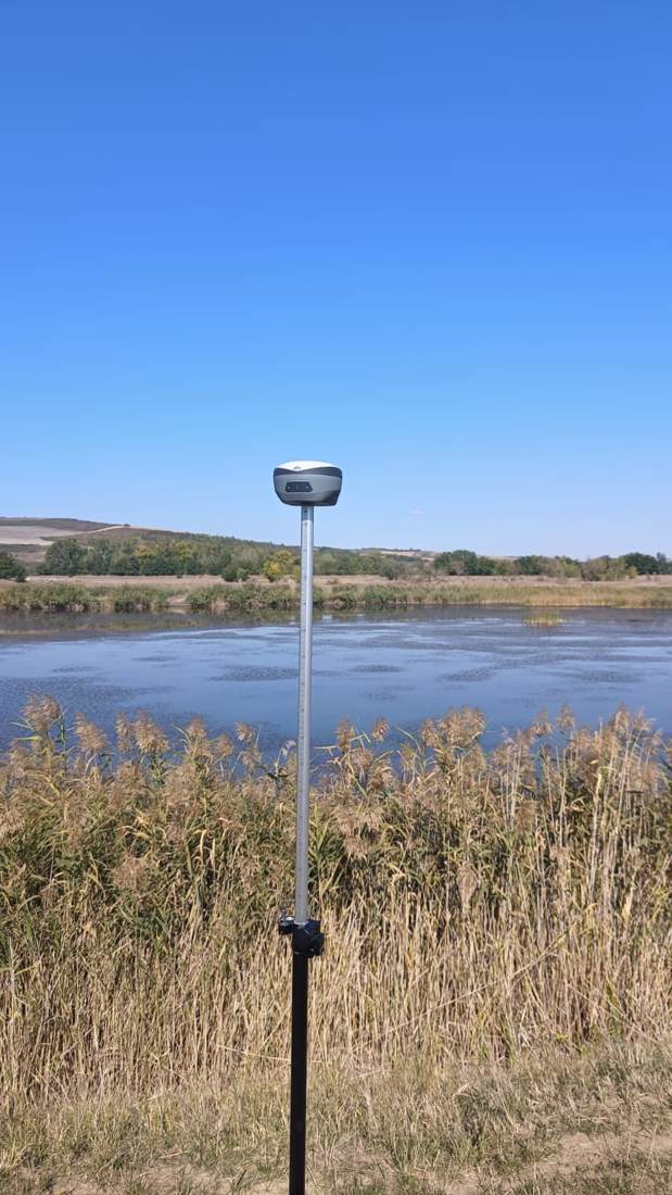 Receptor GNSS montat pe o tijă de măsurare, lângă un lac, într-o zi însorită.