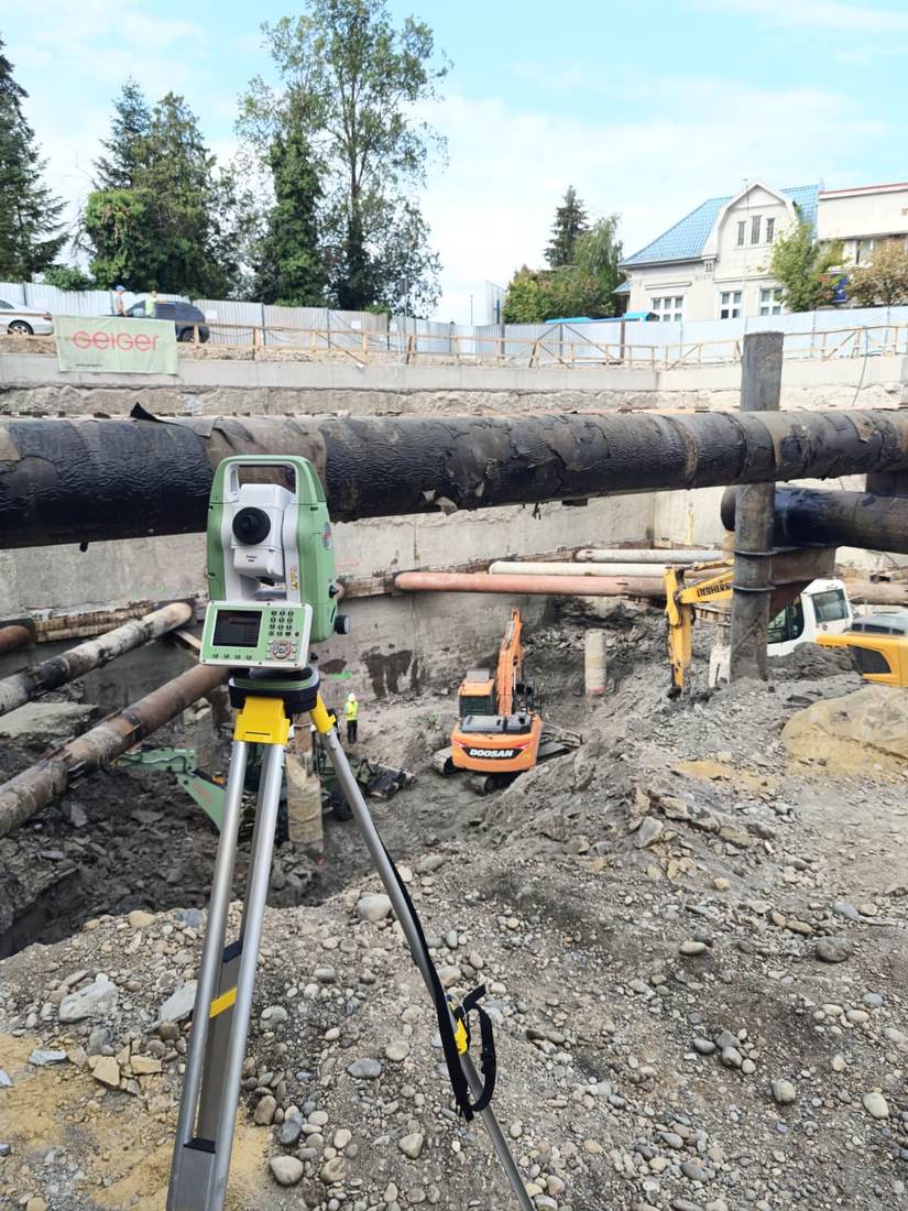 Stație totală poziționată pe șantier, deasupra unei excavații ample cu conducte subterane.