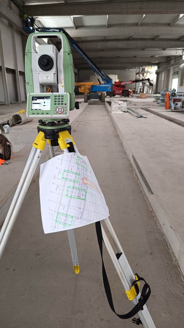 Stație totală amplasată într-o hală industrială, cu un plan tehnic prins de picioarele trepiedului.