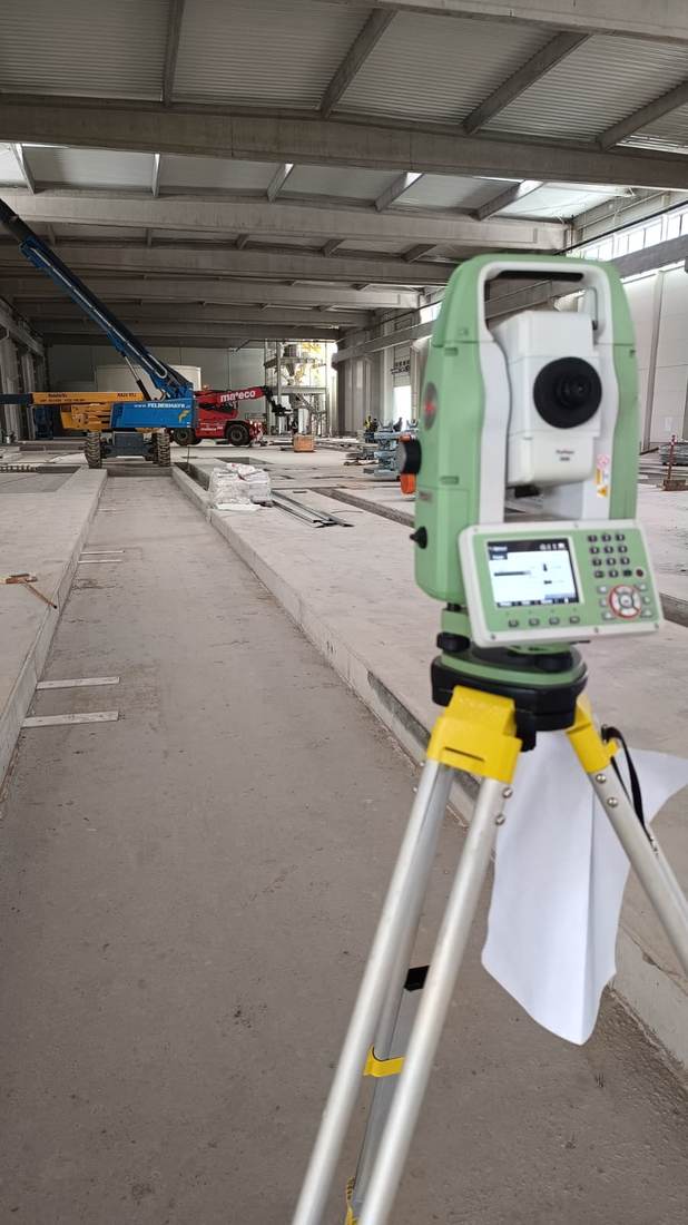 Stație totală în prim-plan, într-o hală industrială cu structură metalică, în construcție.
