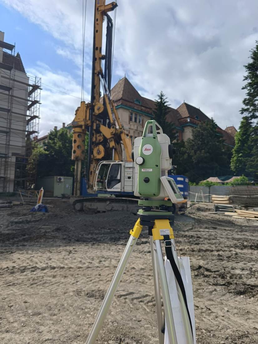 Stație totală Leica, cu un utilaj de foraj pentru piloți în fundal, pe un șantier din centrul orașului.