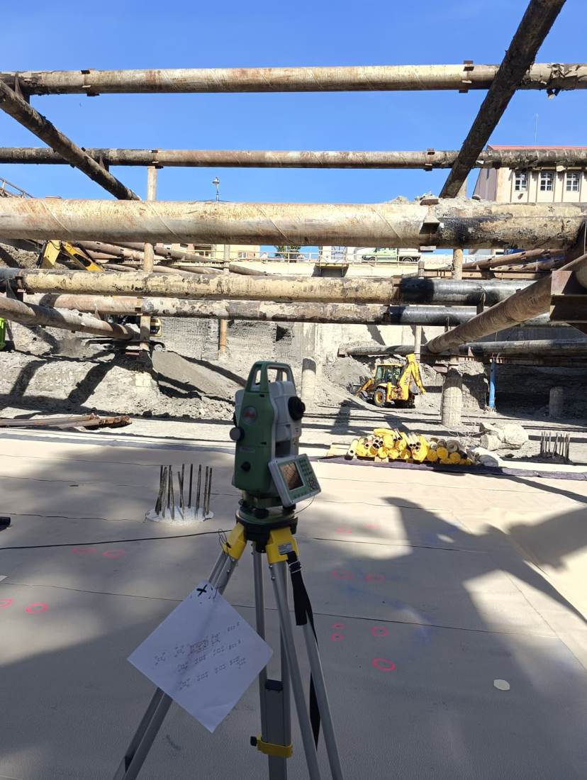 Stație totală Leica pe un șantier de excavare, între conducte și utilaje de lucru.