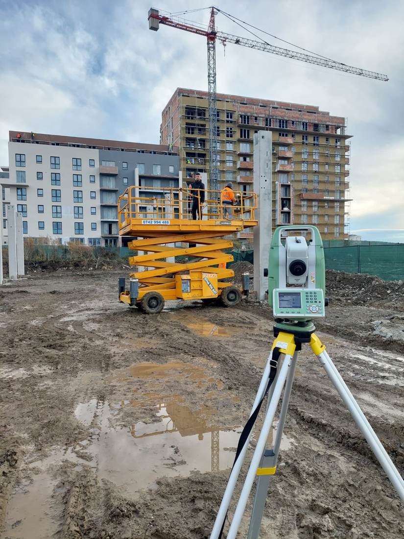 Stație totală pe șantierul unui bloc de locuințe în construcție, cu macara și platformă mobilă.
