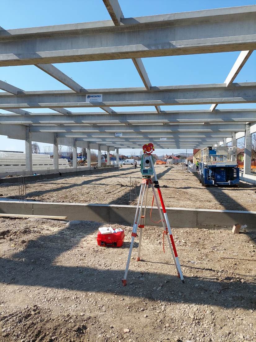 Stație totală sub o structură amplă din beton, în construcție, cu platformă mobilă în fundal.