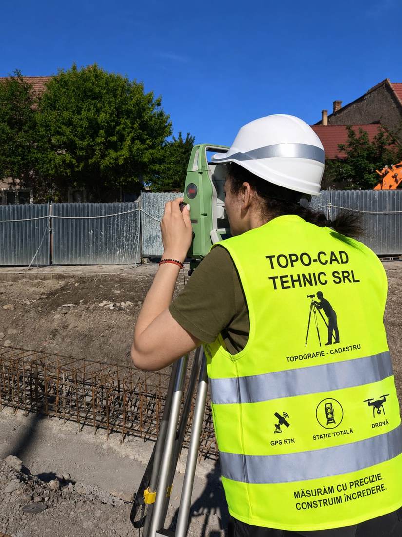 Membru al echipei, echipat cu vestă reflectorizantă și cască de protecție, operând stația totală.